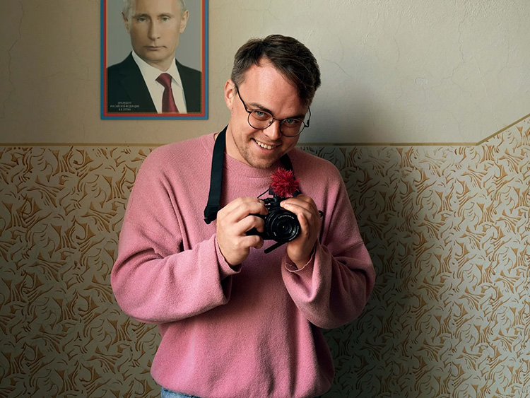 A man in a pink sweatshirt smiles and holds a camera next to a photo of Vladimir Putin hanging on the wall