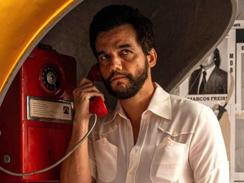 A man with a beard stands beside a mid-20th Century payphone, holding the receiver up to his ear