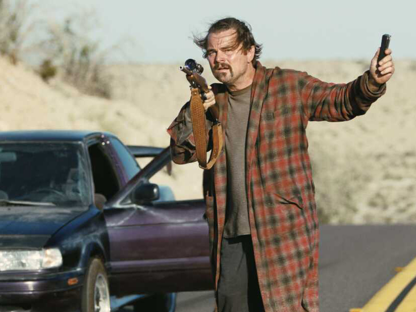 A man in a flannel robe stands beside an older-model sedan in the middle of a desert highway, aiming a rifle with one hand and raising a cylindrical object in the other